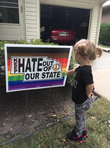 Picture of a little girl with big tails pointing at a yard sign with a rainbow background and a Nebraska state shape with the words 